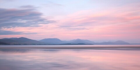 Fototapeta premium A serene landscape photograph captures a tranquil lake reflecting a pastel-colored sky and distant mountains. The water surface is remarkably smooth, mirroring the soft hues of pink, purple