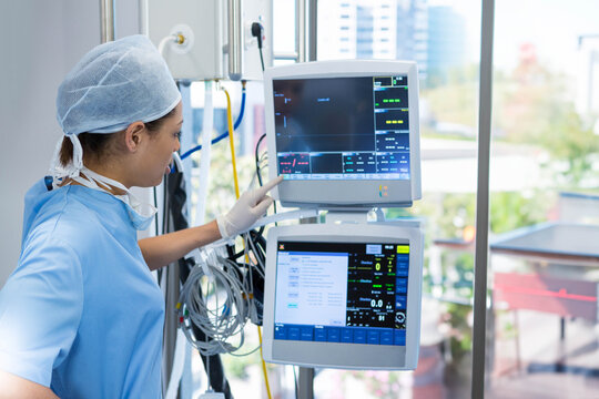 Nurse monitoring patient vital signs on dual screens beside large window in hospital room