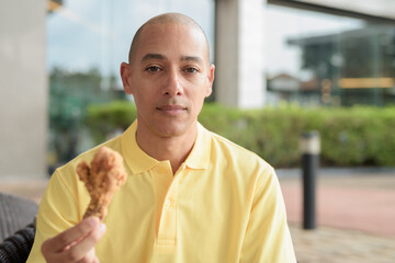 Bald Hispanic man eating fried chicken for lunch during day outdoors