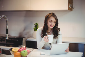 Asian woman sitting at kitchen island holding ceramic mug and looking at tablet near fruit bowl