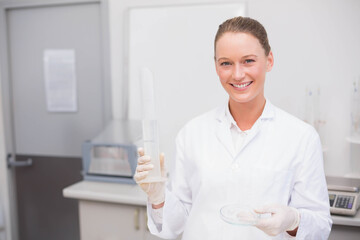 Female lab technician standing at bench wearing coat, gloves holding graduated cylinder, petri dish