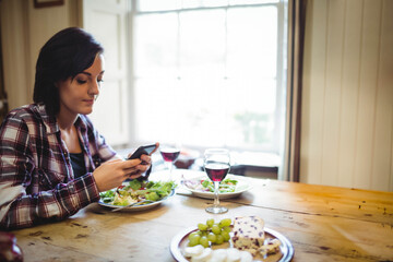 Woman sitting at wooden table by window holding phone above salad, wine glasses, cheese, grapes