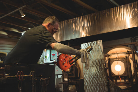 Molten glass glowing red on blowpipe above workshop bench amid furnace, tools, ventilation ducting