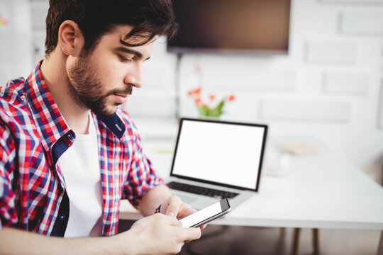 Bearded man scrolling smartphone at desk in home office with laptop, coffee mug, copy space