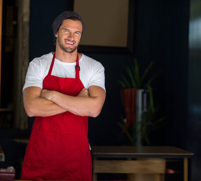 Male barista wearing red apron and knit beanie, crossing arms, smiling behind wooden table at café - Powered by Adobe