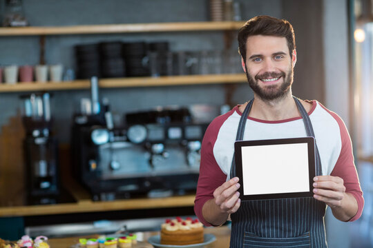 Bearded man barista holding tablet standing in cafe with pastries and espresso machine - Powered by Adobe
