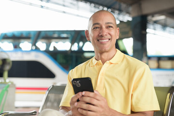 Hispanic bald tourist man waiting for bus and using phone at station platform