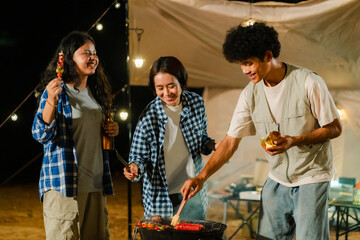 Group of Asian friends enjoying a camping barbecue at night under warm firelight. Woman grills colorful skewers next to warm tent, creating a fun and friendly outdoor dining atmosphere.