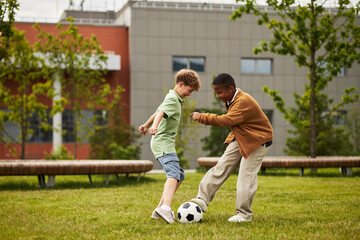 Obraz premium Two boys, one Caucasian and one Black, playing soccer on grass field outside, actively competing for ball, modern building visible in background