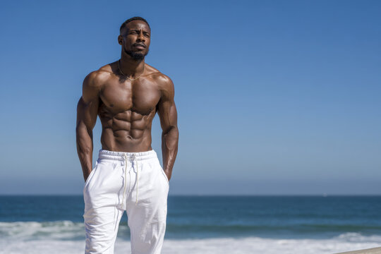Fit muscular man wearing white outfit posing confidently on a beach with ocean in the background