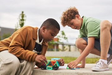 Black boy and Caucasian boy building geometric shapes with colorful magnetic construction toys outdoors, both sitting on concrete surface and focusing on creative play together