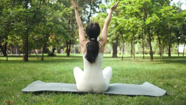 Young fit sporty happy slim Asian woman wearing beige sportswear doing yoga stretching on mat in nature park.