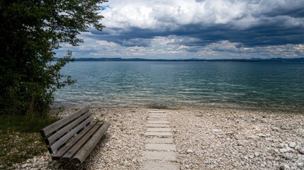 A wooden bench sits on a rocky beach, facing a calm lake and a cloudy sky.
