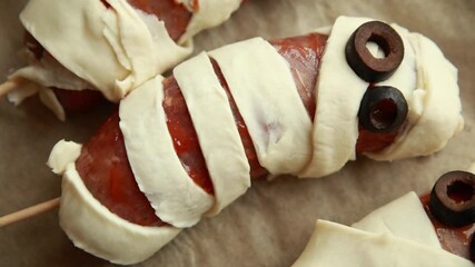 raw Halloween appetizer mummies made from sausages wrapped in puff pastry and decorated with olives. Festive spooky snack idea, ready to be baked for Halloween party food