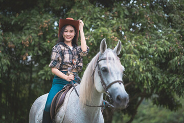 Fototapeta premium Beautiful young Asian woman in cowgirl fashion happily riding a horse at an outdoor training ground in Chiang Mai, Thailand.