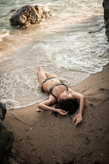 Asian woman in black swimsuit lying on sandy beach at sunset, waves touching her body among stones, summer vacation and tropical seascape concept