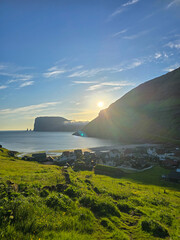 Golden sunlight casts a warm glow over the charming village of Tjornuvik, nestled between lush green hills and the tranquil ocean of the Faroe Islands