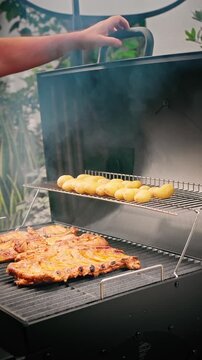Juicy pork ribs grilling on lower rack, small potatoes roasting above inside smoky outdoor barbecue smoker.