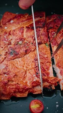 Close-up of juicy grilled pork ribs being sliced with a knife, sprinkled with coarse salt and herbs, served with cherry tomato on black slate.