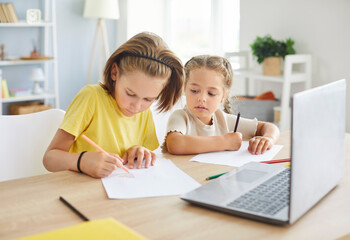 Portrait of two preteen children sitting at table with laptop, drawing pictures with pencils together. Focused kids, cute little brother and sister engaged in creative activities together at home.