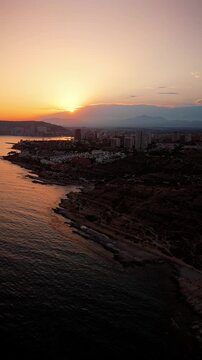 Drone video capturing Spanish coastline and city skyline at sunset, with golden light reflecting on calm sea and rugged shore.