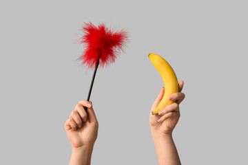Female hands with feather stick from sex shop and banana on grey background, closeup