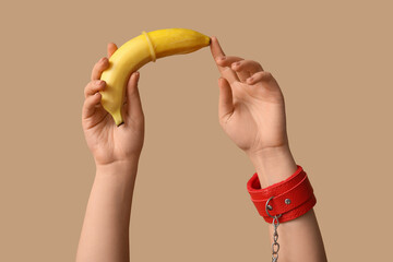 Woman in handcuffs from sex shop holding banana with condom on color background, closeup