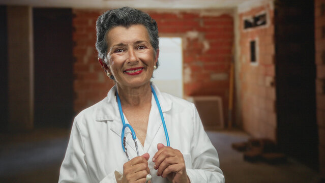 Senior woman doctor with grey hair and stethoscope smiling at an indoor construction site wearing a white coat against a brick wall background.