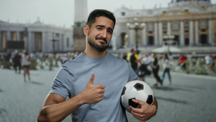 Young man smiling with thumbs up holding soccer ball in vatican's saint peter's square, giving...