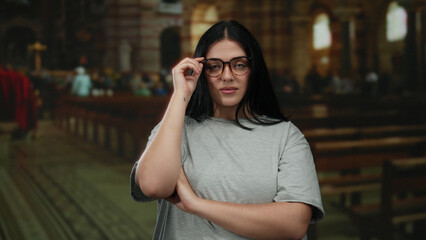 Fototapeta premium Young woman with glasses standing confidently in a church, wearing casual attire and expressing empowerment through her pose in an indoor religious setting.