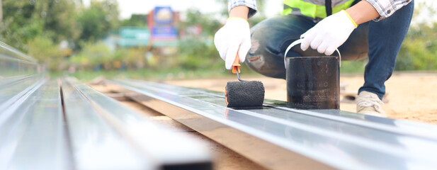 Construction worker painting steel beam with black protective coating using roller brush. Concept...