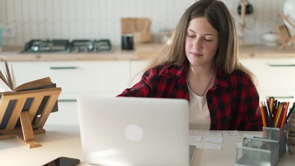 Teenager studying with laptop and study card on kitchen desk, girl focused on homework and online learning, organizing note card, pencil holder beside book, natural light, calm education setting