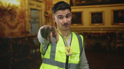 Fototapeta premium Young hispanic man in reflective vest volunteering indoors at museum points with engaging expression.