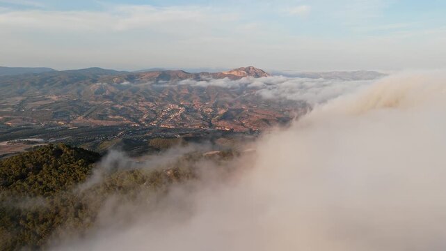 Drone video of Spanish mountains with dense clouds flowing over forested slopes, revealing peaks under clear blue sky.