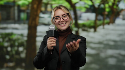 Woman reporter holding microphone outdoors in a park making a gesture as if inviting someone to come closer while smiling confidently.