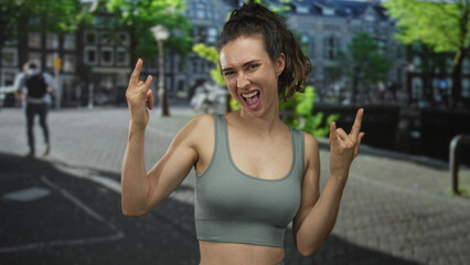 Sunlit urban woman in tanktop hands making rock horns gesture in bustling street; rock metal...
