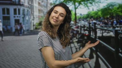 Young hispanic woman in striped shirt holds out both hands in a welcoming gesture on a busy street; confidence.