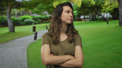Fototapeta premium Woman in olive shirt crosses arms and looks sideways in a green park with a paved path under shaded trees; rejection.