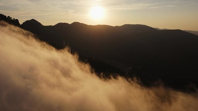 Drone video of mountain peaks surrounded by golden clouds at sunset, with warm light illuminating the misty slopes in Spain.