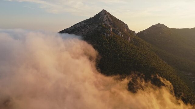 Drone video over Spanish mountain peak covered with forest, with golden sunset light illuminating drifting clouds in the foreground.