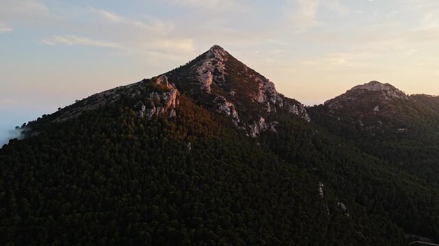 Aerial drone video of a forest-covered mountain peak in Spain at sunrise, with warm sunlight casting golden tones on rocky slopes.