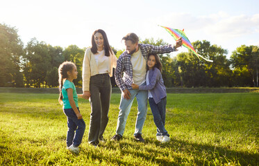 Fototapeta premium Summer green park, happy family standing smiling warmly, holding preparing colorful kite for flying, feeling joyful and connected embracing on sunny weekend day, playing game together for fun