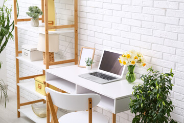 Shelving unit and workplace with bouquet of narcissus flowers and laptop near white brick wall in office