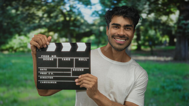 Man smiling broadly while holding clapboard and lifting clapper stick in forest under green foliage; joy.