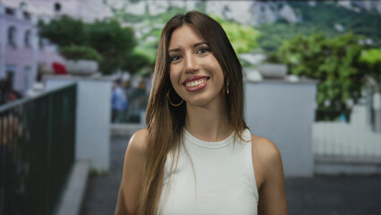 Woman wearing hoop earrings and white sleeveless top smiling with bare shoulders on a busy street;...