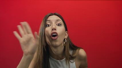 Woman raises bare hand palm forward in a red studio with hoop earrings, lips parted and wide eyes; curiosity.
