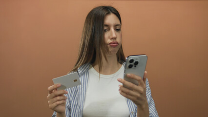 Woman holding creditcard and smartphone against orange studio wall with focused gaze; uncertainty decision.