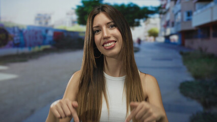 Obraz premium Young hispanic woman in white tank top points fingers downward on sunny city street; friendly invitation.