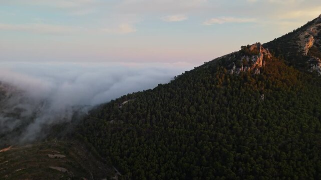 Aerial drone video of a forested mountain peak in Spain, bathed in warm sunlight, emerging above dense clouds at sunrise.