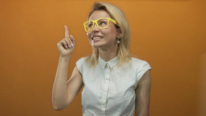 Young blonde woman with yellow eyeglasses pointing finger upward with a smile in orange studio; inspiration.